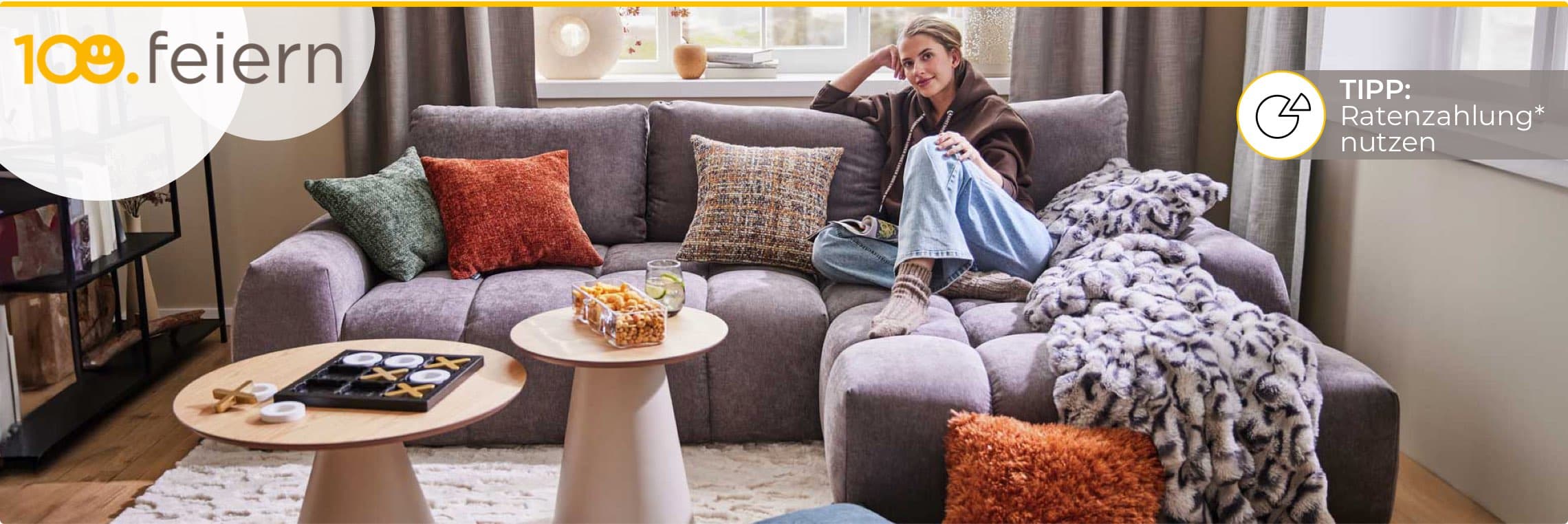 100. feiern – Tipp: Ratenzahlung nutzen – Person entspannt auf grauem Sofa mit bunten Kissen und Snacks auf Couchtischen