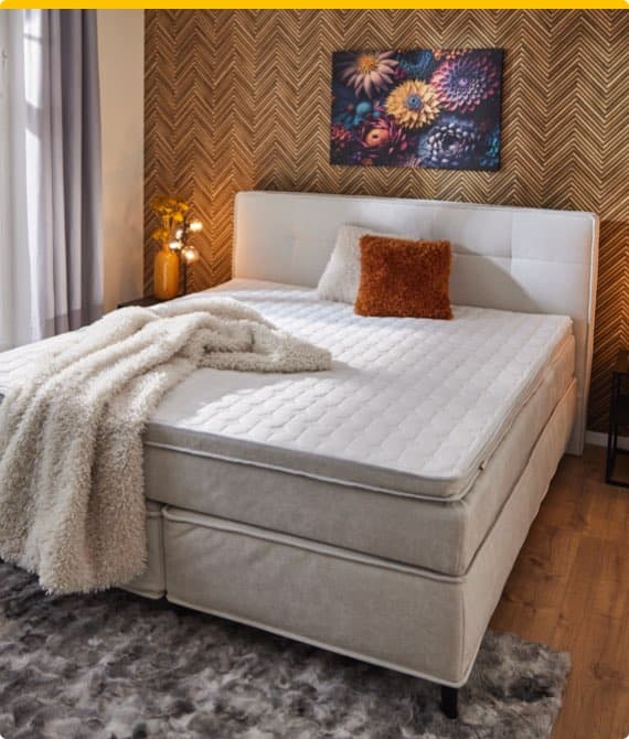 Gemütliches Schlafzimmer mit Doppelbett, weißer Tagesdecke und Kissen, buntes Blumenbild an der Wand