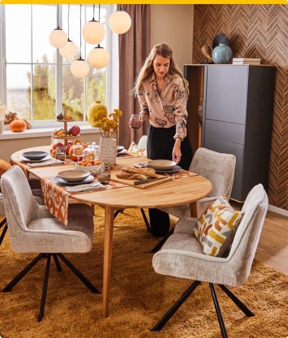 Frau deckt einen abgerundeten Esstisch, dekoriert mit Herbstmotiven, in einem gemütlichen Esszimmer mit modernen hellgrauen Stühlen.