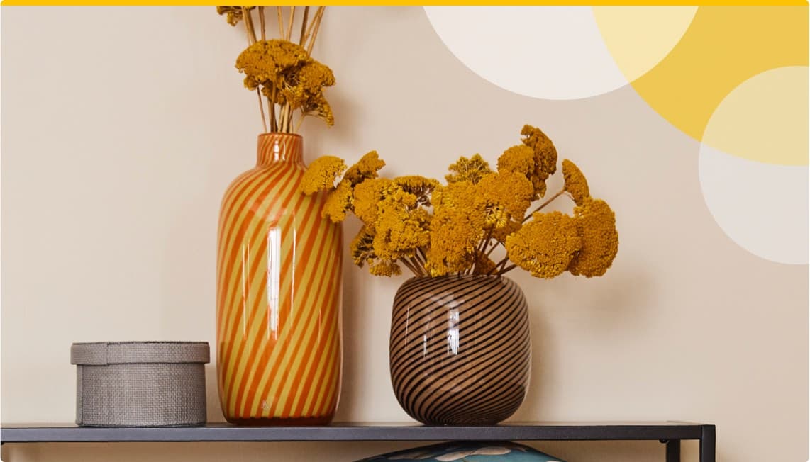 Zwei Vasen mit gelben getrockneten Blumen, eine große gestreifte orange Vase und eine kleinere gestreifte braune Vase, daneben eine graue Box