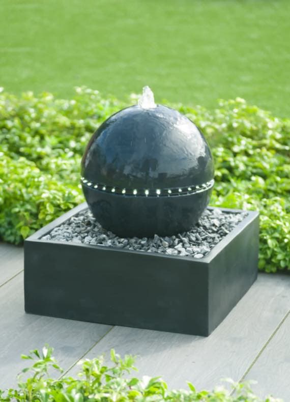 Schwarzer Kugelbrunnen mit LED-Beleuchtung auf Kiesbett, umgeben von grünen Pflanzen im Garten.
