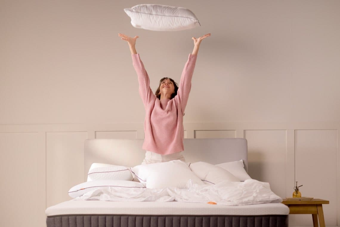 Frau in rosa Pullover wirft Kissen in die Luft, sitzt auf einem ungemachten Bett.