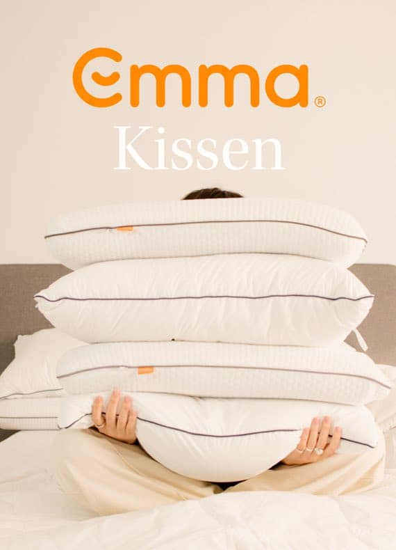 Emma Kissen – Person hält vier weiße Kissen auf dem Bett, Kopf hinter den Kissen verborgen.