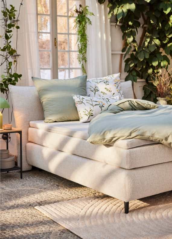 Gemütliches Bett mit grüner Bettwäsche und Kissen in lichtdurchflutetem Raum mit Pflanzen und großen Fenstern.