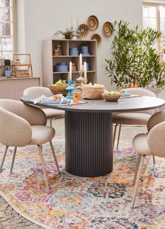 Runder Esstisch mit Stühlen, gedeckt mit Essen, vor einem Regal mit Geschirr und Pflanzen im Hintergrund.