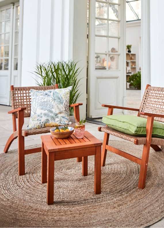 Zwei Holzstühle mit Kissen und ein kleiner Tisch auf einem runden Teppich vor einer weißen Tür mit Glasfenstern.