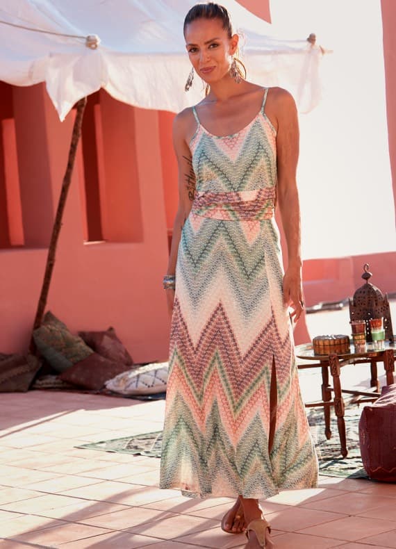 Frau im langen Kleid mit Zickzackmuster steht auf Terrasse vor rosa Wand und orientalischer Sitz- und Deko-Ecke