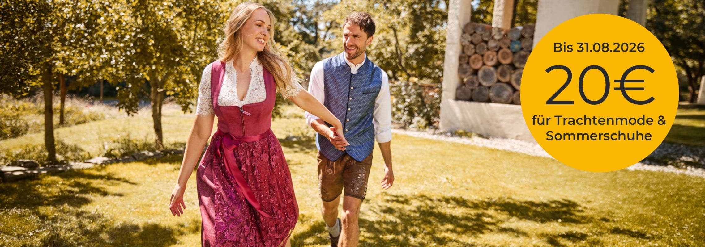 Bis 31.08.2026 20€ für Trachtenmode & Sommerschuhe – Paar in Tracht hält sich im sonnigen Park an den Händen