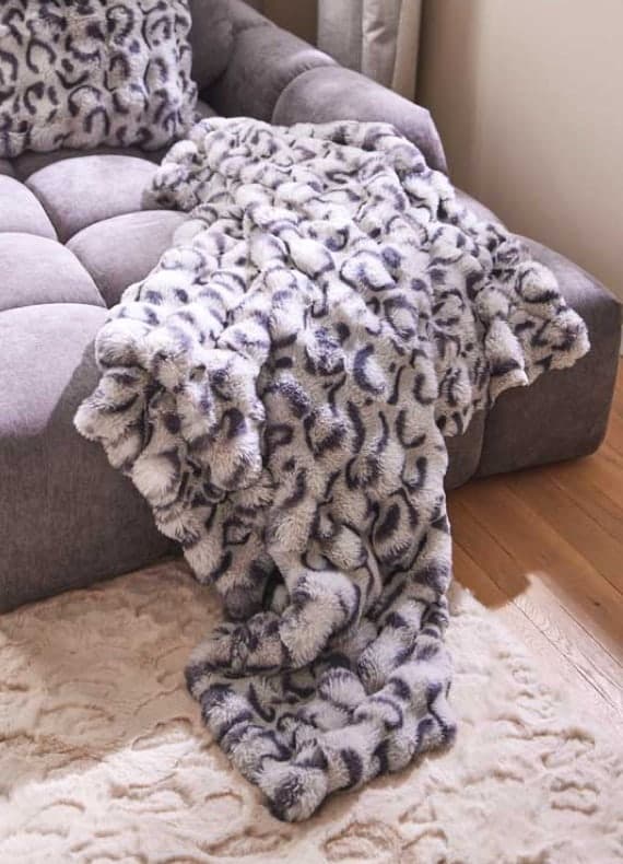 Grau gemusterte Decke mit passendem Kissen liegt locker über einem grauen Sofa und reicht bis auf den Teppich