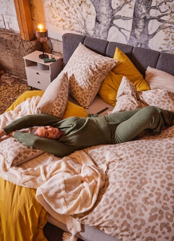 Frau in grüner Loungekleidung liegt ausgestreckt auf Bett mit gemusterter Bettwäsche und gelben Kissen in gemütlichem Schlafzimmer