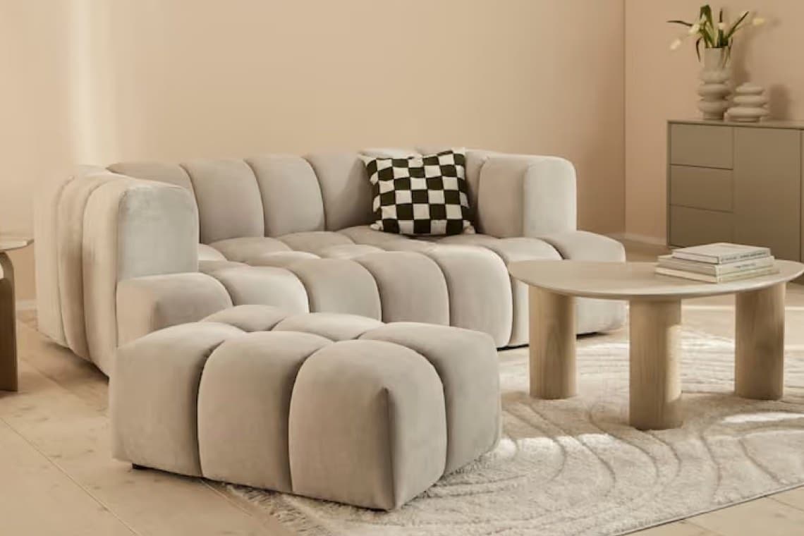 Beiges Sofa mit passendem Hocker, schwarz-weiß kariertes Kissen, runder Couchtisch und Teppich im hellen Wohnzimmer