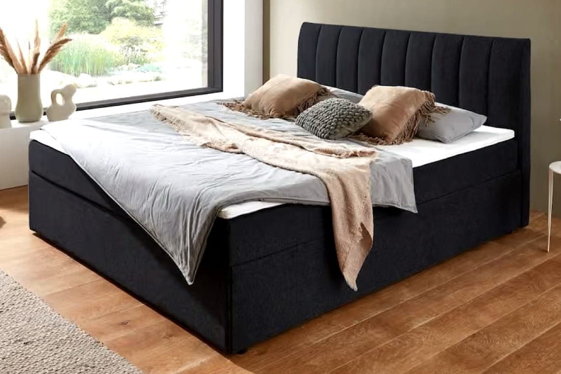 Dunkles Boxspringbett mit hohem Kopfteil, grauer Bettwäsche und Kissen, neben großem Fenster im Schlafzimmer