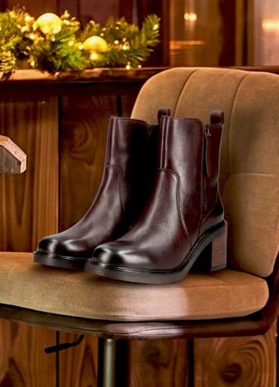 Dunkelbraune Stiefeletten mit Blockabsatz stehen auf einem Polsterstuhl, im Hintergrund weihnachtliche Deko mit Lichtern