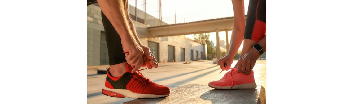Zwei Personen binden ihre Sportschuhe, eine in roten und eine in pinken Sneakers, bei Sonnenuntergang in urbanem Umfeld.