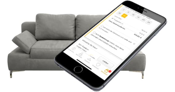 Smartphone zeigt Buchungsseite, dahinter graues Sofa mit Kissen und Armlehne
