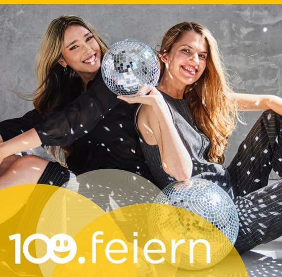 100 feiern – Zwei Frauen sitzen mit Disco-Kugeln am Boden, sie tragen festliche Kleidung und lachen