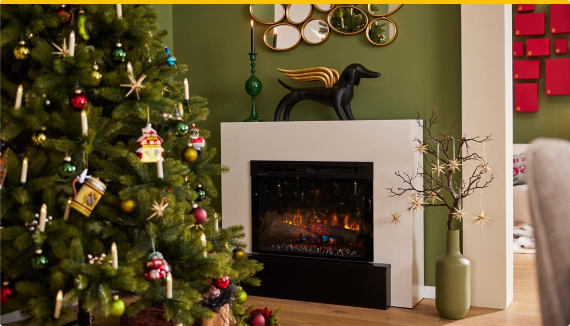 Weihnachtlich geschmückter Baum neben Kamin, darüber Dekofigur und Kerzenhalter, Spiegel an der grünen Wand, Vase mit Zweigen und Sternen