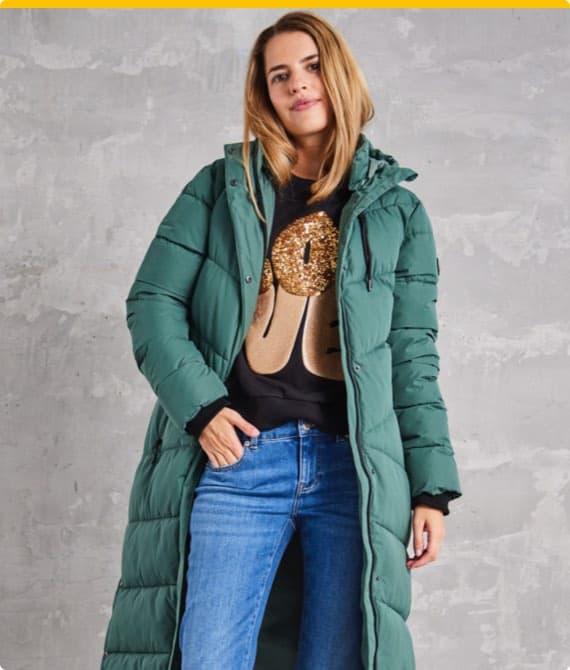 Frau in grünem Wintermantel und schwarzem Pullover mit goldener Grafik hält Hände in den Taschen ihrer blauen Jeans