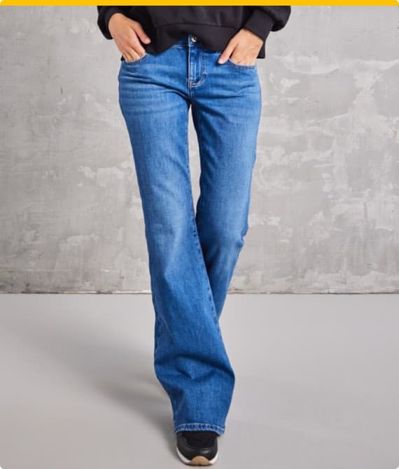 Person in blauer Schlagjeans und schwarzen Schuhen vor grauem Hintergrund. Hände in den Taschen.