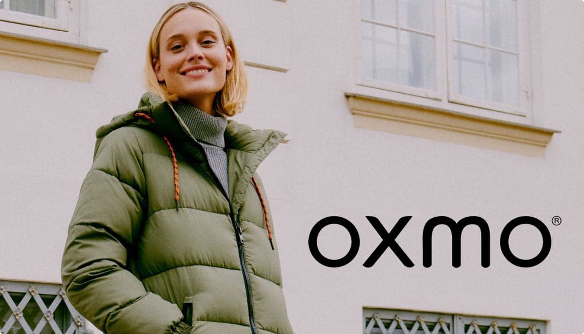 oxmo – Person trägt eine grüne Daunenjacke vor einem Gebäude mit Fenstern und Gitter.
