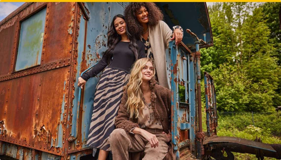 Drei Frauen stehen und sitzen vor einem verrosteten, blauen Eisenbahnwaggon in einem grünen Wald.