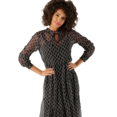 Frau trägt ein schwarzes Kleid mit langen Ärmeln und geometrischem Muster. Hände in die Hüften gestützt.