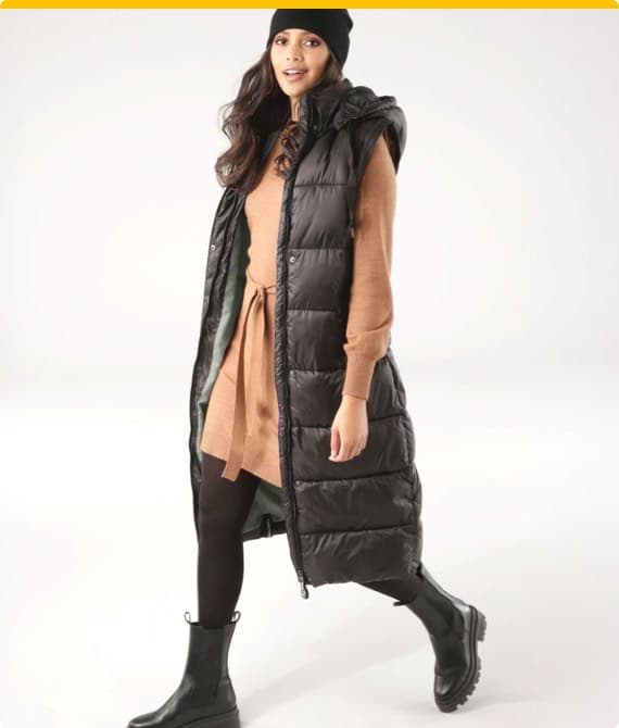 Frau in braunem Pulloverkleid und langer schwarzer Steppweste geht, trägt schwarze Stiefel und Mütze.