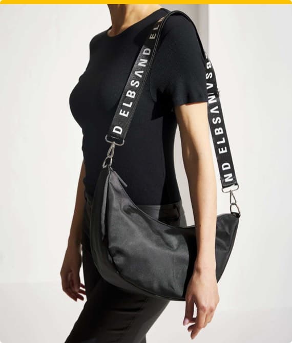 Person mit schwarzer Tasche, breite Träger mit Aufschrift "ELBSAND", trägt schwarzes Shirt.