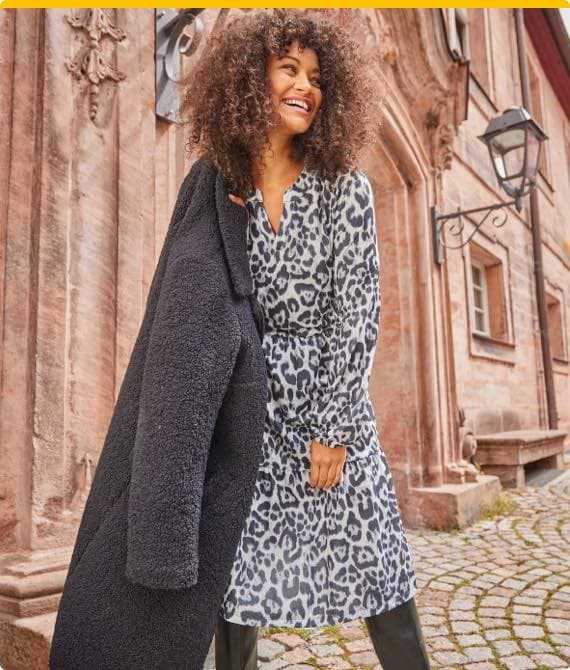 Frau in grauem Leopardenkleid hält einen schwarzen Mantel über der Schulter, steht vor einem historischen Gebäude.