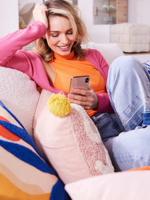 Frau in orangefarbenem Oberteil und pinker Strickjacke hält ein Smartphone und sitzt auf einem Sofa.