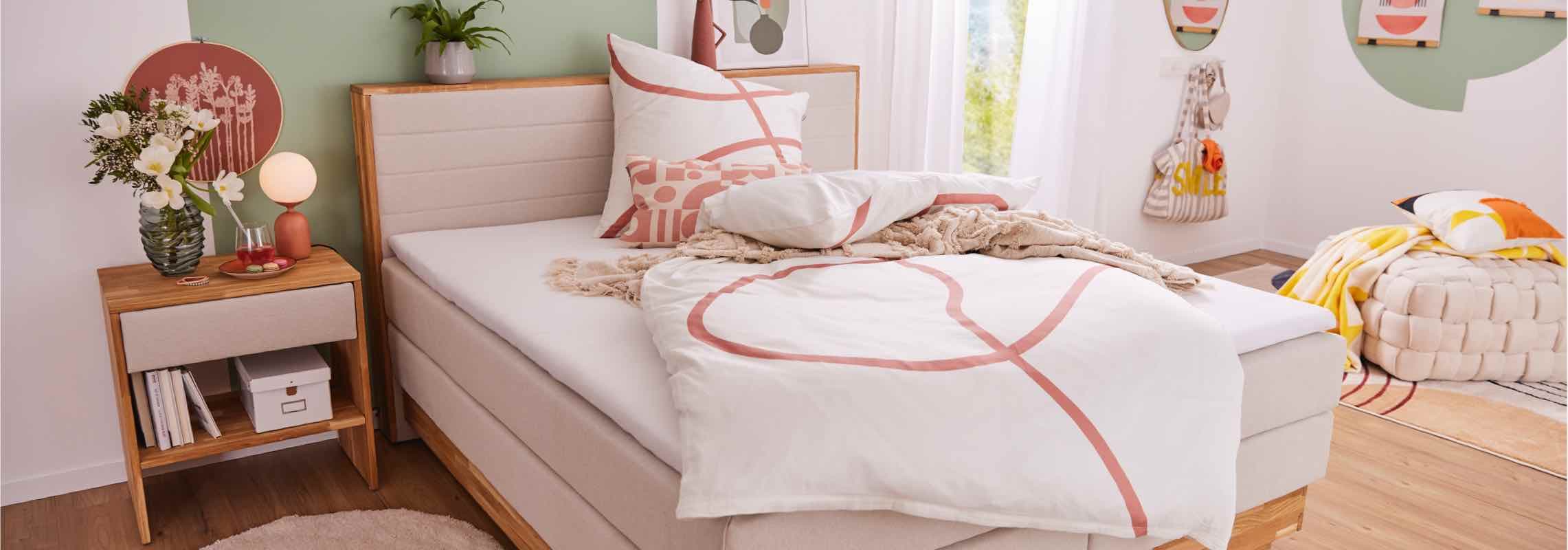 Modernes Schlafzimmer mit beigem Bett, Nachttisch, Blumen, Lampe und dekorativer Bettwäsche in Weiß-Rosa.