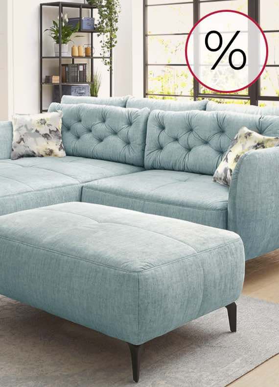 Helles Wohnzimmer mit hellblauer Polsterecke und Hocker, Regal im Hintergrund