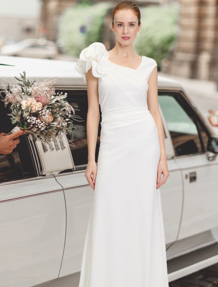 Braut in weißem Kleid mit Blumenschmuck steht vor einem weißen Auto