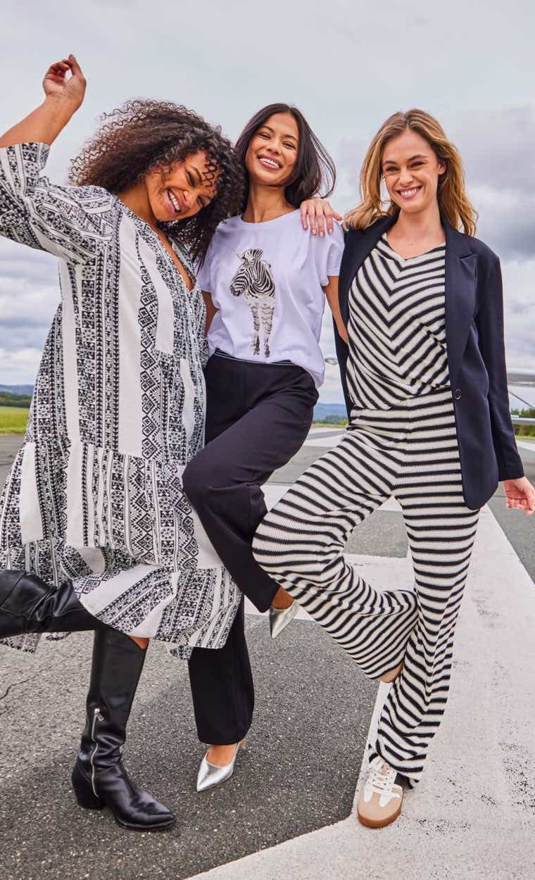Drei Frauen in schwarz-weiß gemusterter Kleidung tanzen fröhlich auf einer Straße.