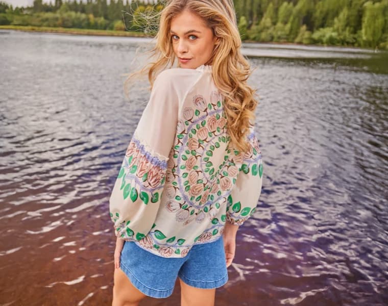 Frau mit blonder Locken trägt bunt gemusterte Bluse und Jeansshorts, steht am Seeufer mit Blick auf das Wasser und den Wald.