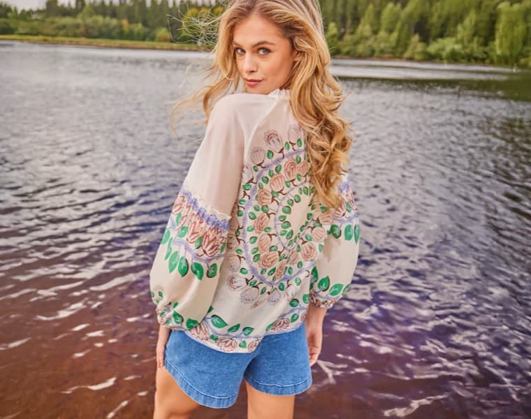 Frau am See trägt eine gemusterte Bluse und Jeansshorts, Rückenansicht mit lockigem blondem Haar.