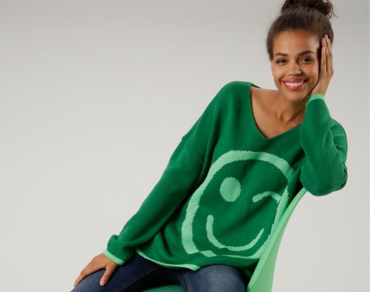 Frau in grünem Pullover mit großem Smiley-Design, sitzt auf einem grünen Stuhl und stützt ihren Kopf mit der Hand.