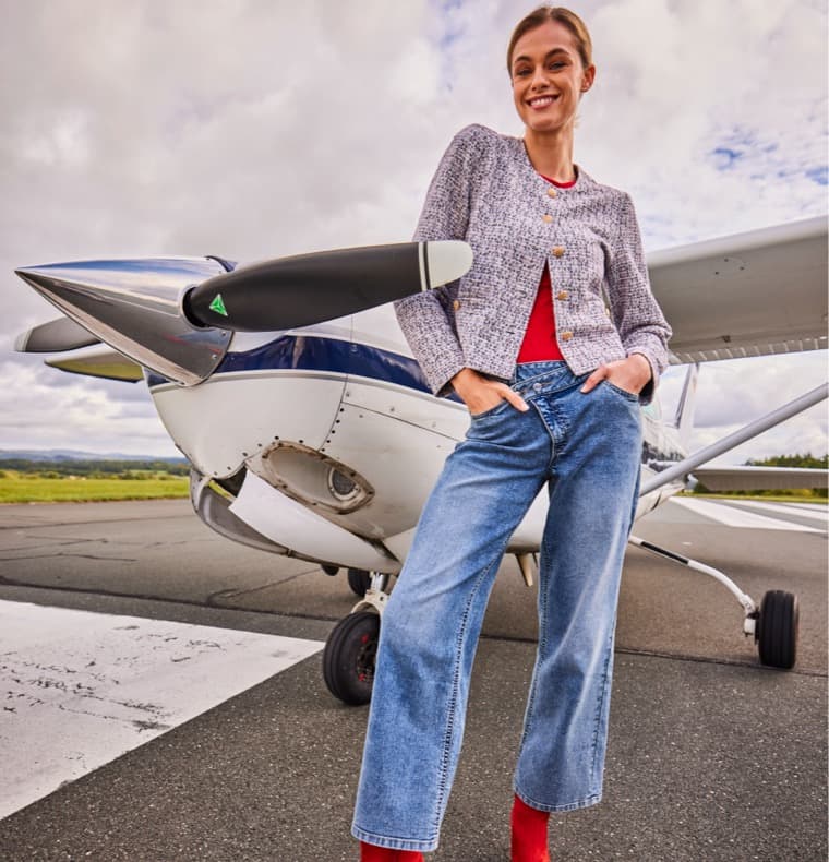 Frau in rotem Shirt, grauer Jacke und blauer Jeans steht vor einem Flugzeug auf dem Rollfeld.