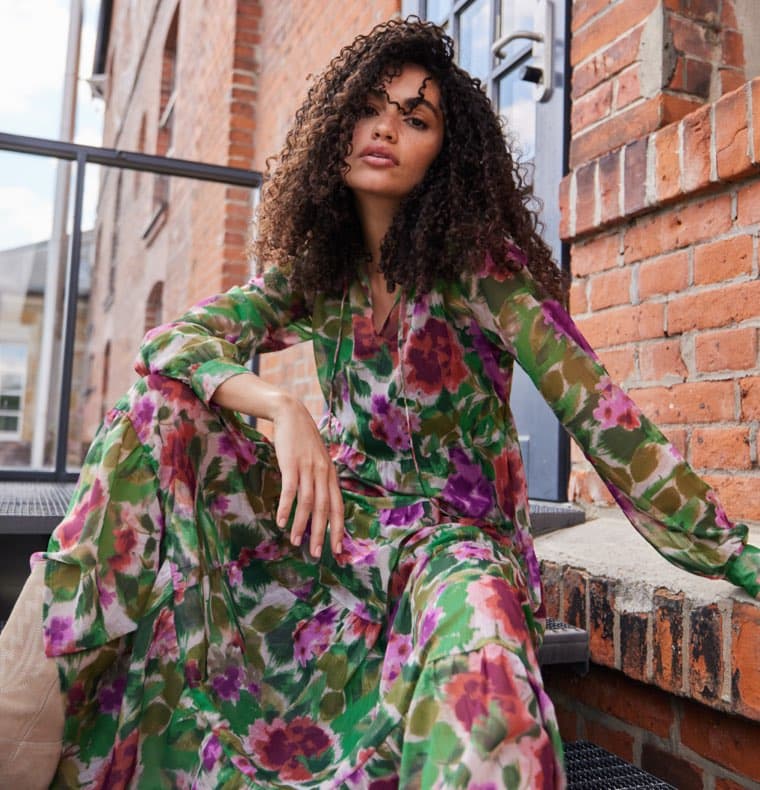 Frau in langem, buntem Blumenkleid sitzt vor Backsteinwand auf Treppe.