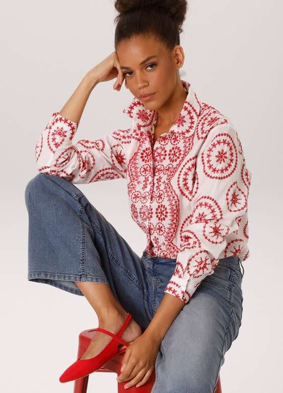 Frau in weißer Bluse mit rotem Muster und blauer Jeans sitzt auf rotem Hocker.