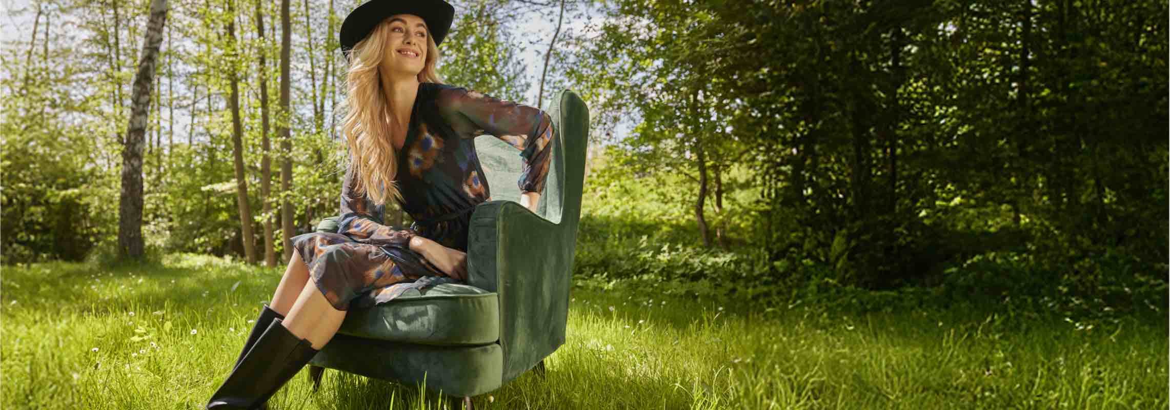 Frau mit Hut sitzt in einem grünen Sessel auf einer Wiese im Wald, trägt ein gemustertes Kleid und schwarze Stiefel.