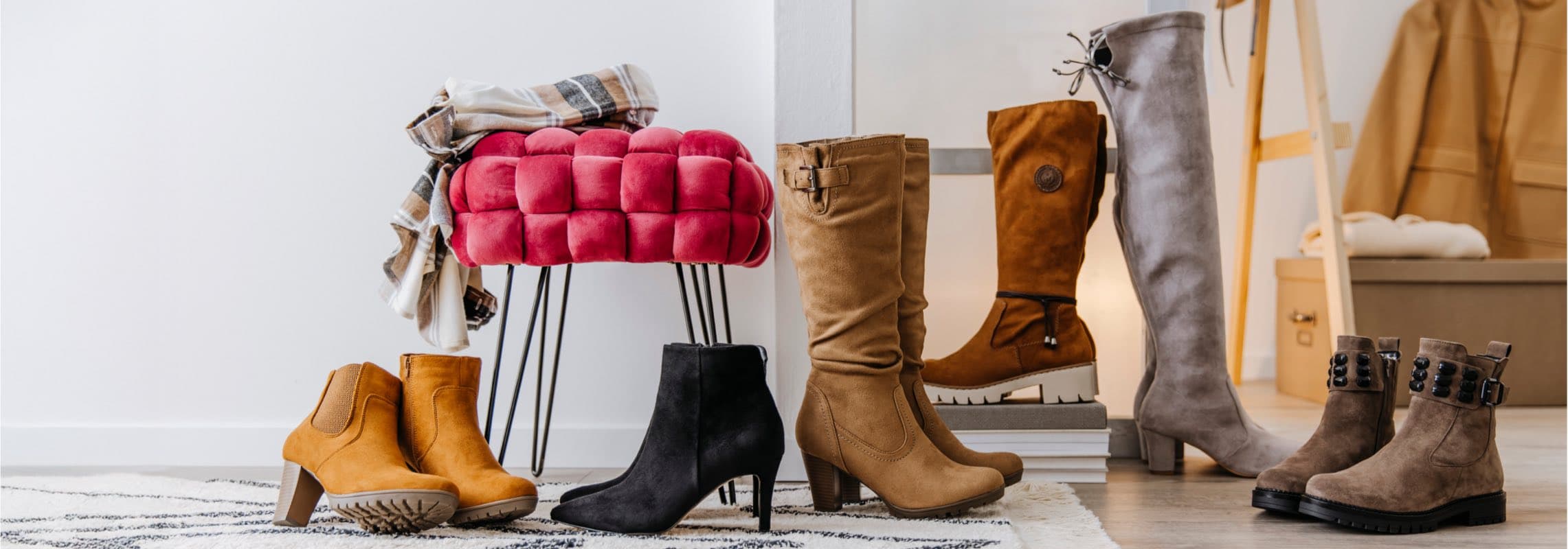 Verschiedene Stiefel und Ankle Boots in Braun, Schwarz und Grau vor einem roten Hocker und einem beigen Mantel.