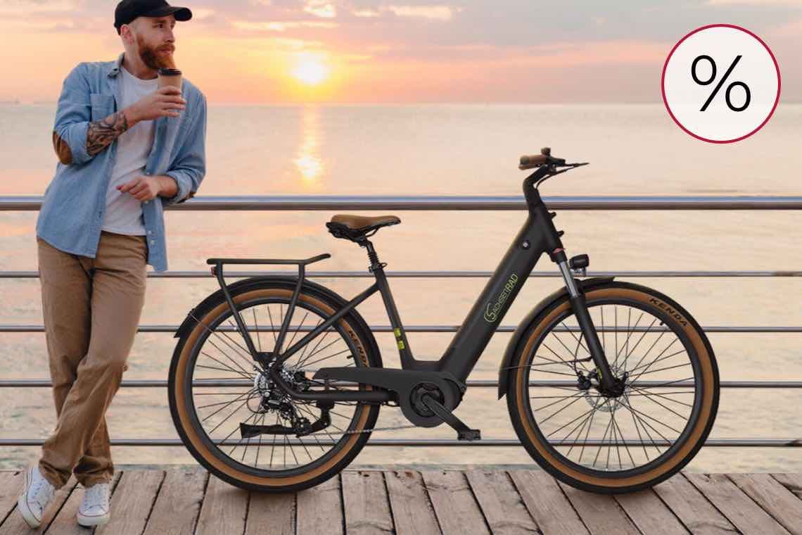 % – Mann lehnt am Geländer, hält einen Kaffee, neben ihm steht ein modernes Fahrrad, im Hintergrund Sonnenuntergang am Meer