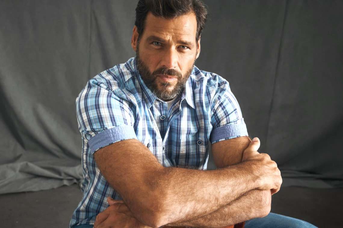 Mann in kariertem Hemd sitzt mit verschränkten Armen vor einem grauen Hintergrund. Gesicht ist unscharf.