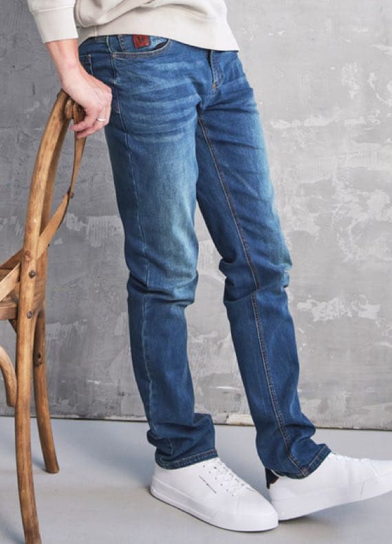 Person trägt blaue Jeans und weiße Sneakers, lehnt sich an einen Holzstuhl vor grauer Wand.