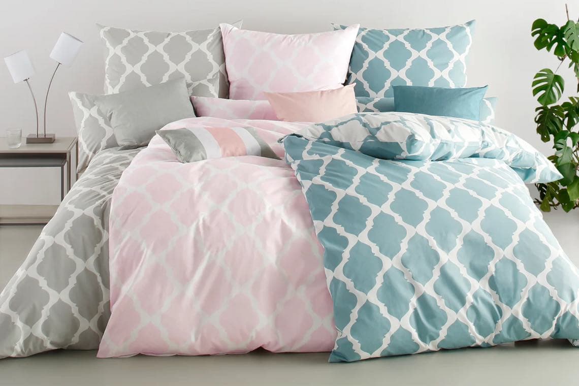 Bett mit gemustertem Bettzeug in Rosa, Grau und Blau, umgeben von dekorativen Kissen und Nachttisch mit Lampen.