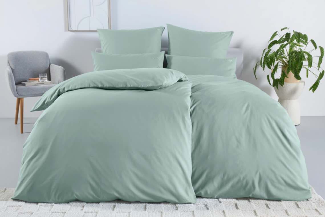 Mintgrüne Bettwäsche auf einem Bett, daneben ein grauer Sessel und eine grüne Zimmerpflanze.