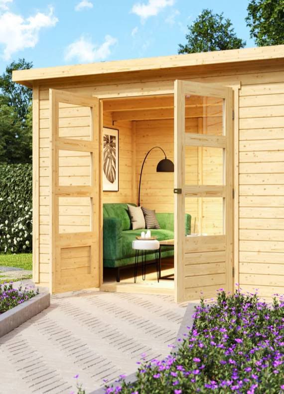 Offene Holztür eines Gartenhauses mit grünem Sofa und schwarzer Stehlampe im Inneren, umgeben von Blumen.