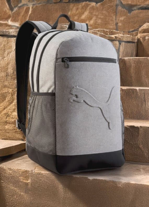 Grauer Rucksack mit Puma-Logo auf Steintreppe, Fronttasche mit Reißverschluss, seitliche Netztaschen.