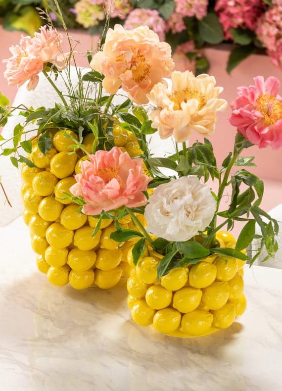 Zwei gelbe Vasen aus kleinen Kugeln, gefüllt mit rosa und weißen Blumen, stehen auf einem Tisch.